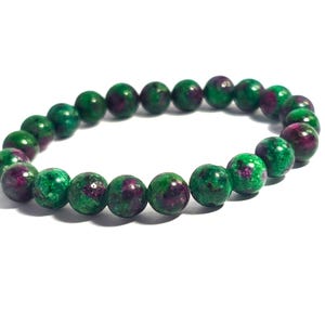 May include: A beaded bracelet featuring round, polished beads in shades of green and maroon. The bracelet is strung together, creating a circular shape. The beads have a speckled appearance, adding visual interest. The bracelet is set against a white background.