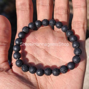 Puede incluir: Un brazalete de cuentas de piedra de lava negra. El brazalete está hecho de cuentas redondas y porosas. El texto "ShreekrishnagemsShop" es visible en la imagen. El brazalete se muestra en una mano.