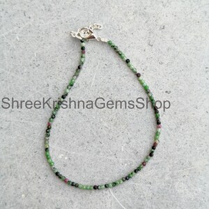 May include: A delicate beaded bracelet featuring small, faceted green and dark red gemstone beads arranged in a continuous circular strand. The bracelet includes a silver-toned metal lobster claw clasp and a small extender chain at the top.