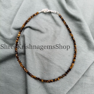 May include: A necklace made of small, round tiger's eye beads in brown and gold hues. The necklace has a silver clasp and is displayed on a grey surface. The text "Shree KrishnagemsShop" is visible in the image.