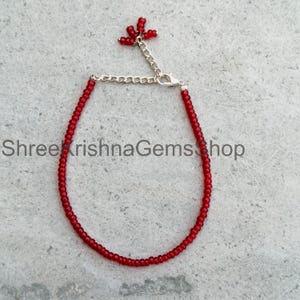 May include: A delicate red beaded bracelet featuring small, uniform red beads strung on a thin wire. The bracelet includes a silver-toned metal chain extender with a small cluster of red beads at the end. The text ShreeKrishnaGemsShop is visible.