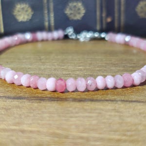 Könnte beinhalten: Ein zartes Armband aus rosa Edelsteinen. Das Armband besteht aus kleinen, facettierten, runden Perlen in verschiedenen Rosatönen, die mit einem silbernen Verschluss aufgefädelt sind. Das Armband wird auf einer Holzoberfläche präsentiert.