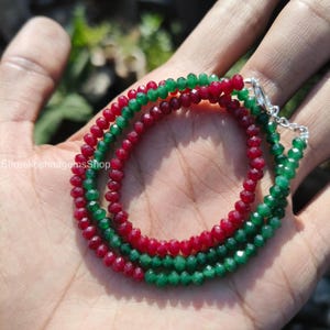 Pode incluir: Um close-up de três fios de colares de contas. Os colares são feitos de pequenas contas facetadas em cores vermelha e verde alternadas. Os colares são conectados com um fecho prateado. Os colares são exibidos na mão de uma pessoa.