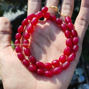 Könnte beinhalten: Eine zweireihige Halskette mit ovalen, tiefroten Perlen. Die Perlen sind zu einer Halskette mit einem goldfarbenen Verschluss aufgereiht. Die Halskette wird auf einer Hand präsentiert.
