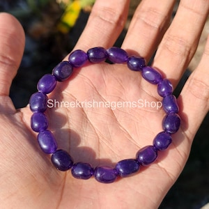 May include: A bracelet made of polished, oval-shaped, deep purple beads. The beads are strung together to form a circular shape. The bracelet is held in a hand, with the text "Shreekrishnagemsshop" visible.