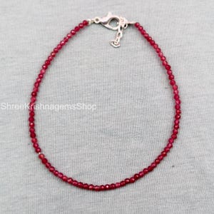 May include: A delicate red gemstone bracelet with a silver clasp. The bracelet is made of small, round, red beads strung together. The clasp is a lobster claw style. The bracelet is displayed on a gray surface.