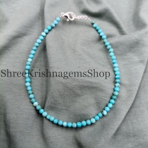 May include: A delicate turquoise beaded bracelet with a silver clasp. The small, round beads are a vibrant blue-green color. The bracelet is displayed on a gray fabric surface. The text "ShreeKrishnagemsShop" is visible.