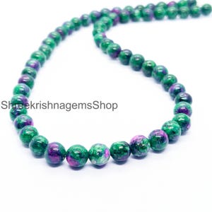 Peut inclure: Un collier de perles rondes vertes et violettes. Les perles sont enfilées ensemble, formant une ligne incurvée. Le texte "ShreekrishnagemsShop" est visible sur le côté gauche de l'image.