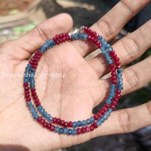 Pode incluir: Colar de duas voltas com contas alternadas de pedras preciosas vermelhas e azul-esverdeadas. O colar é preso com um fecho prateado. As pequenas contas facetadas criam um efeito brilhante. O colar é exibido em um fundo neutro.