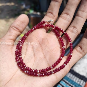 May include: A necklace of small, faceted, deep red gemstones interspersed with silver-coloured beads. The necklace is shown on a hand, highlighting its colour and design. The clasp is visible at the top.