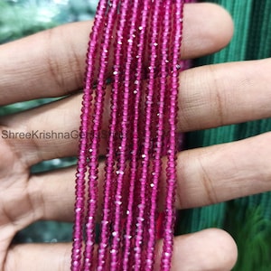 Könnte beinhalten: Nahaufnahme von mehreren Strängen kleiner, facettierter, tiefrosa Perlen. Die Perlen sind miteinander aufgereiht und erzeugen einen strukturierten, schimmernden Effekt. Die Perlen werden in einer Hand gehalten, mit einem grünen und tealfarbenen Hintergrund.