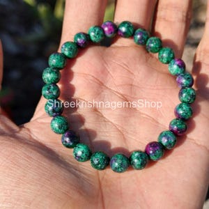 Puede incluir: Una pulsera de cuentas con piedras preciosas verdes y moradas. Las cuentas redondas presentan una mezcla de tonos verdes oscuros y morados vibrantes. El texto "Shreekrishnagemsshop" es visible.