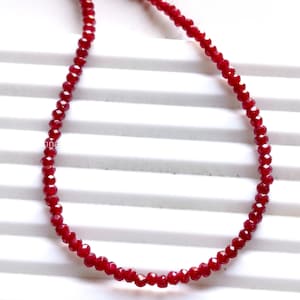 Può includere: Una collana composta da piccole perline rosse sfaccettate. Le perline sono infilate insieme per formare un filo continuo. La collana ha un aspetto scintillante, che riflette la luce. La collana è presentata su uno sfondo bianco.
