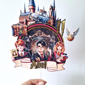 Può includere: Un colorato cake topper con illustrazioni di personaggi di una serie fantasy. Il design include un castello, un'auto volante e un boccino d'oro. I personaggi tengono in mano una bacchetta, libri e una carta. Il nome "Abel" è visualizzato.