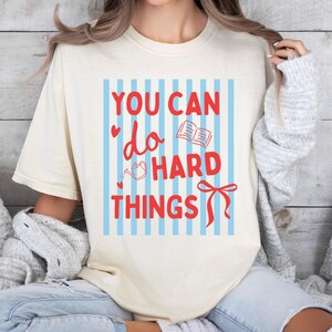 May include: A cream-colored short-sleeve t-shirt featuring a graphic with vertical light blue stripes and the red text "YOU CAN do HARD THINGS." The design includes small red heart icons, a line-art drawing of a book, a watering can, and a red bow.