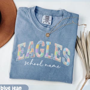 Puede incluir: Una camiseta azul vaquero con la palabra "EAGLES" en un diseño de patchwork colorido. Debajo de la palabra "EAGLES" está el texto "school name" en una fuente de escritura. Un collar de oro con un colgante descansa sobre la camiseta.