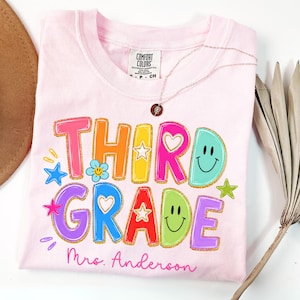May include: A light pink t-shirt featuring the text "THIRD GRADE" in colorful, bold letters with gold outlines. The design includes smiley faces, stars, and hearts. Below the text, the name "Mrs. Anderson" is printed in pink script.