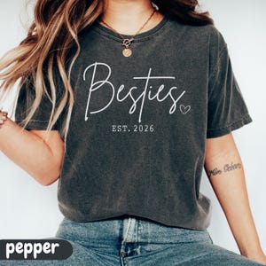 May include: A dark gray t-shirt with the word "Besties" in white script, accompanied by "EST. 2026" and a small heart. The shirt is a casual style, suitable for everyday wear.