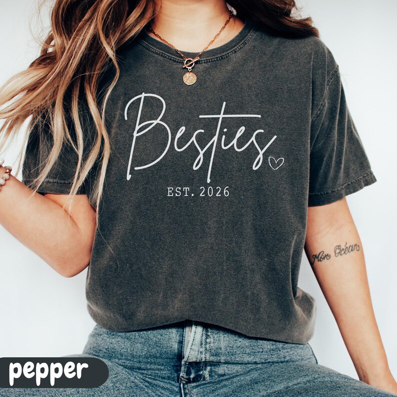 May include: A dark gray t-shirt with the word "Besties" in white script, accompanied by "EST. 2026" and a small heart. The shirt is a casual style, suitable for everyday wear.