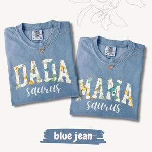 May include: Two blue jean-colored t-shirts with the words "DADA saurus" and "MAMA saurus" in white block letters with dinosaur patterns. Each shirt has a gold necklace. The shirts are on a white background.