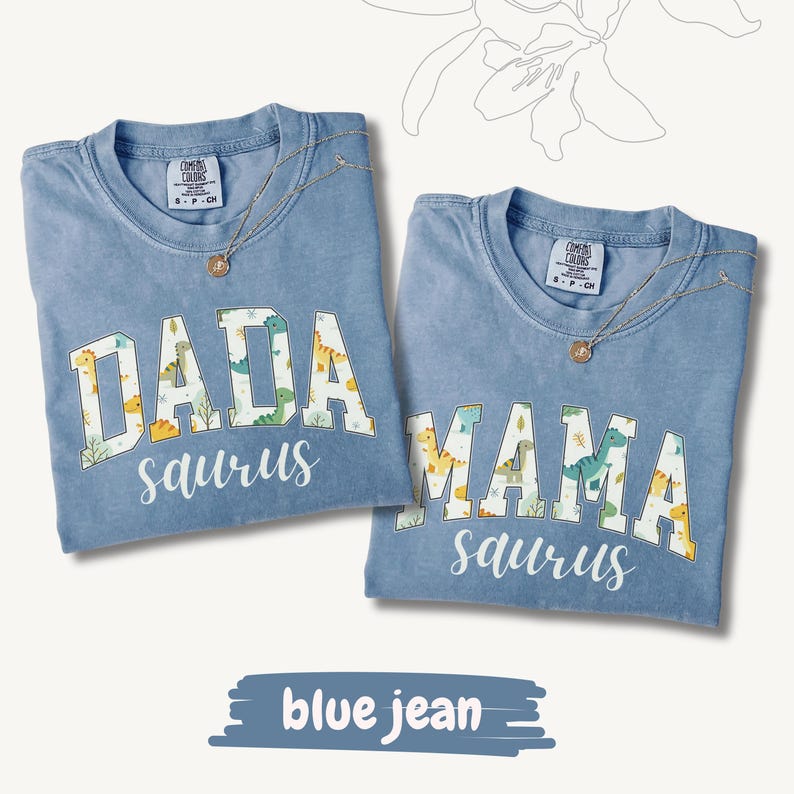 May include: Two blue jean-colored t-shirts with the words "DADA saurus" and "MAMA saurus" in white block letters with dinosaur patterns. Each shirt has a gold necklace. The shirts are on a white background.