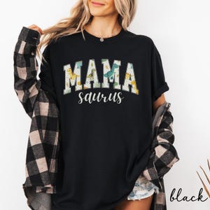 May include: Black t-shirt with the text "MAMA saurus" in white and a dinosaur design. The shirt has a crew neck and short sleeves. The shirt is paired with a black and white plaid shirt.