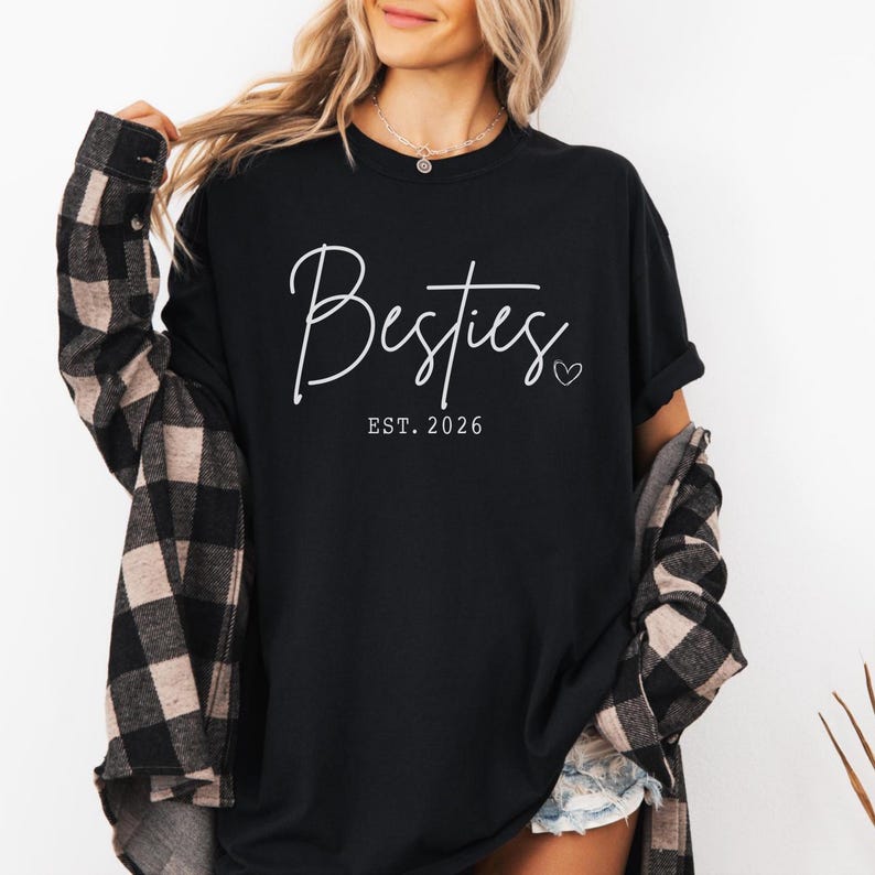 May include: Black t-shirt with the word "Besties" in a cursive font, with "EST. 2026" below it. A small heart is next to the word "Besties". The person is wearing a black and tan plaid shirt over the t-shirt.