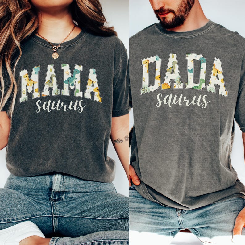 May include: Two gray t-shirts with the words "MAMA saurus" and "DADA saurus" printed on them in white, with a dinosaur pattern inside the letters. The shirts are worn by two people, one wearing blue jeans.
