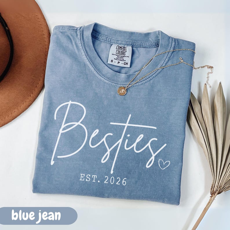 blue jean sweatshirt with “Besties EST. 2026” design in white script font, folded with necklace and jeans for a cozy flatlay