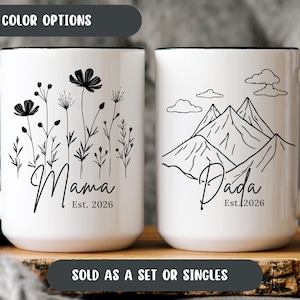 May include: Two white ceramic mugs with black handles and rims. One mug features a floral design with the word "Mama" and "Est. 2026." The other mug has a mountain design with the word "Dada" and "Est. 2026." The text "SOLD AS A SET OR SINGLES" is also visible.