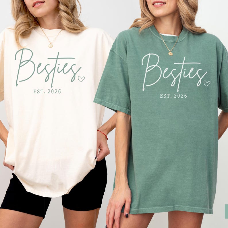 May include: Two t-shirts, one cream and one sage green, each with the word "Besties" in a script font, a small heart, and "EST. 2026" printed below. The shirts are short-sleeved and appear to be made of a soft material.