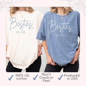 May include: Two t-shirts, one cream and one blue, with the word "Besties" and "EST. 2026" printed on them. The shirts are made of 100% US cotton and produced in the USA. The text is in a script font with a small heart.
