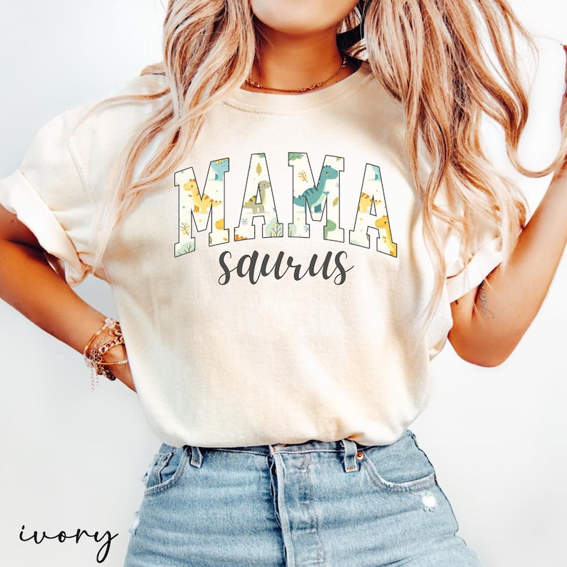 May include: Ivory-colored t-shirt with the words "MAMA saurus" in a playful font. The word "MAMA" is in a green and blue block letter style, with dinosaur and leaf patterns. The word "saurus" is in a cursive font.