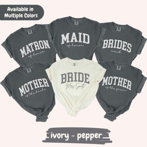 May include: A collection of gray and ivory t-shirts with text for a bridal party. The shirts display phrases like "Matron of Honor," "Maid of Honor," "Bridesmaid," "Mother of the Bride," and "Mother of the Groom." The ivory shirt says "Bride" with a name.