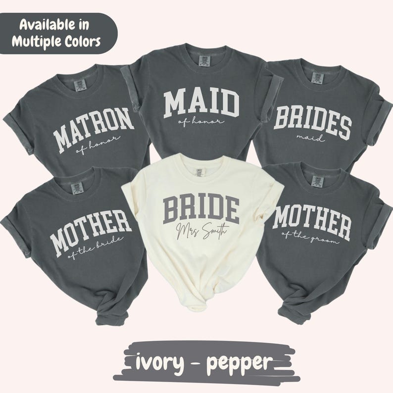 May include: A collection of gray and ivory t-shirts with text for a bridal party. The shirts display phrases like "Matron of Honor," "Maid of Honor," "Bridesmaid," "Mother of the Bride," and "Mother of the Groom." The ivory shirt says "Bride" with a name.