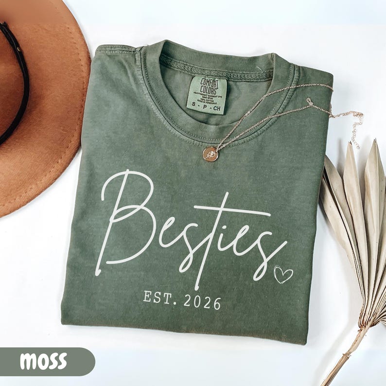 May include: A moss green t-shirt with the word "Besties" in white script, the year "EST. 2026", and a small heart. A gold necklace with a pendant rests on the shirt. A brown hat and dried plant are in the background.