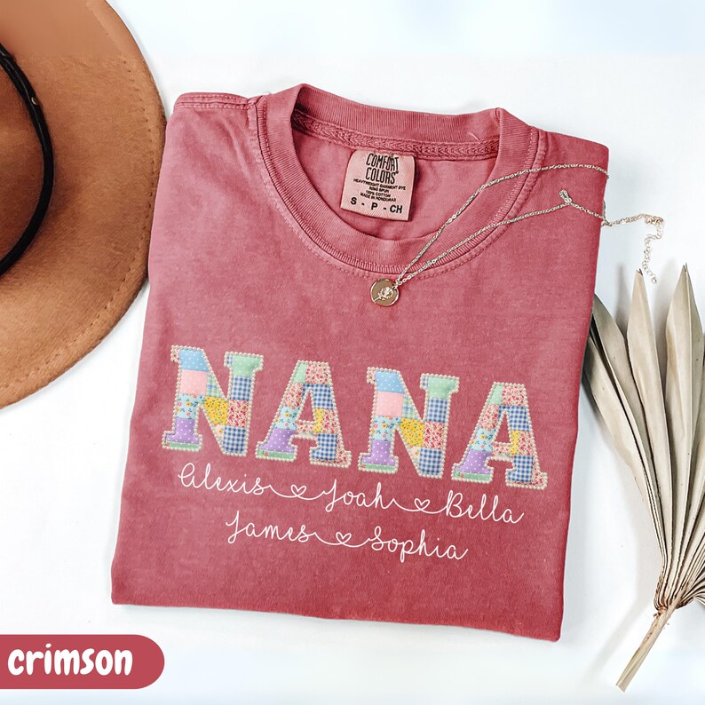 Peut inclure: T-shirt de couleur cramoisie avec le mot "NANA" en lettres patchwork. En dessous, les noms Alexis, Joah, Bella, James et Sophia sont inscrits. Un collier argent&eacute; avec un pendentif est pos&eacute; sur le t-shirt. Un chapeau marron est visible en arri&egrave;re-plan.
