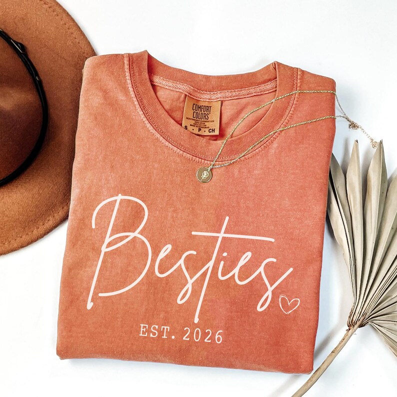 May include: A coral-colored Comfort Colors t-shirt with the word "Besties" in white script, a small heart, and "EST. 2026". A gold necklace with a pendant and a brown hat are next to the shirt.