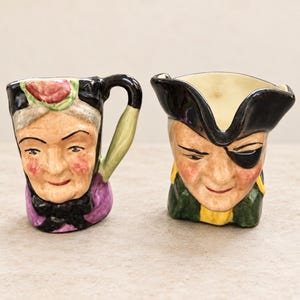 Può includere: Due brocche in ceramica a forma di personaggio. La brocca a sinistra raffigura una persona anziana con cuffia rosa, fiocco nero e abiti viola. Quella a destra raffigura un pirata con tricorno nero, benda sull'occhio e giacca verde e gialla.