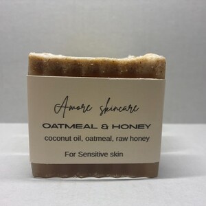 May include: A bar of oatmeal and honey soap from Amore Skincare. The soap is wrapped in a light tan label with the brand name and product details. The ingredients include coconut oil, oatmeal, and raw honey. The label also states the soap is for sensitive skin.