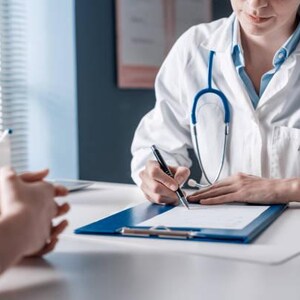 May include: A doctor in a white coat with a stethoscope is writing on a clipboard. The doctor is at a desk with a laptop, facing a patient. The patient's hands are clasped. The scene suggests a medical consultation.