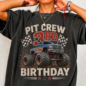 May include: A charcoal gray t-shirt featuring a graphic of a blue monster truck with orange flames, flanked by two checkered racing flags. The text reads PIT CREW 3RD BIRTHDAY in distressed white and red lettering with three small stars below.