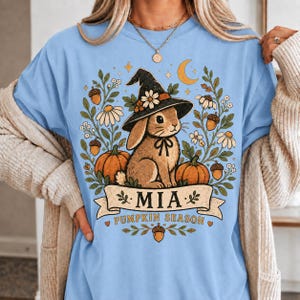 May include: A light blue t-shirt featuring a graphic of a brown rabbit wearing a witch hat, surrounded by pumpkins, acorns, and daisies. Below the rabbit, a banner displays the name MIA and the text PUMPKIN SEASON in dark brown lettering.