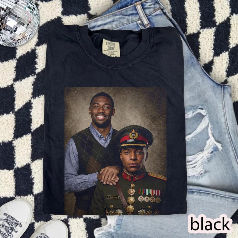 May include: A black t-shirt featuring a graphic print of two individuals. One person wears a light blue button-down shirt and a brown argyle sweater vest. The other person is dressed in a formal military uniform with medals and a peaked cap.