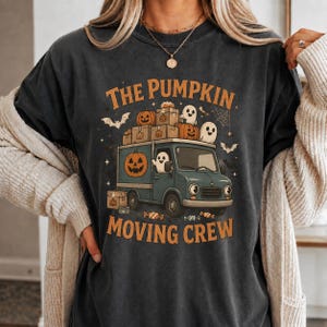 May include: A charcoal gray t-shirt featuring a graphic of a blue delivery truck loaded with pumpkins and ghosts. The text reads THE PUMPKIN MOVING CREW. The design includes small bats, a spider web, and candy illustrations surrounding the vehicle.