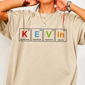 Peut inclure: Un t-shirt beige à manches courtes arborant un motif graphique du prénom KEVIN épelé avec des éléments du tableau périodique. Les lettres K, E, V et In sont affichées en rouge, bleu, vert et orange avec leurs numéros et noms atomiques.