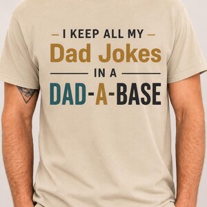 May include: A beige short-sleeve t-shirt featuring the text "I KEEP ALL MY Dad Jokes IN A DAD-A-BASE" printed in black, gold, and teal. The shirt has a crew neckline and a standard fit, with the graphic centred on the chest area of the garment.