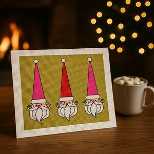 May include: A framed Christmas-themed artwork featuring three Santa Claus faces with pink and red hats, white beards, and mustaches. The artwork is set against a gold background with a cream-colored border. A mug of marshmallows sits nearby.
