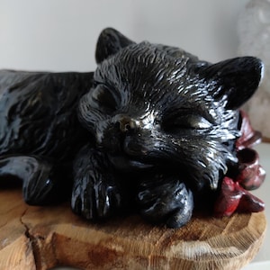 Peut inclure: Sculpture d'un chat noir endormi sur une base en bois. Le chat a les yeux fermés et semble paisible. Des touches de rouge ornent la tête du chat. La sculpture présente une finition texturée et brillante.