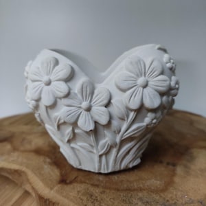 Pode incluir: Um vaso branco em forma de coração com detalhes florais em relevo. O vaso apresenta grandes flores semelhantes a margaridas e flores menores, com folhas e caules. O vaso é feito de um material fosco e de cor clara e está sobre uma superfície de madeira.